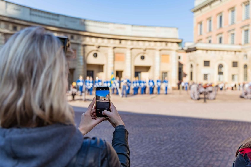 A person taking a photo of a group of people in blue uniforms standing in a plaza with classical architecture in the background. 5
