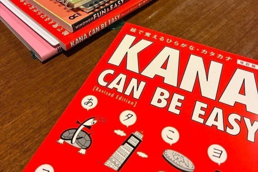 A red book titled 'KANA CAN BE EASY' with Japanese text and illustrations on a wooden surface. 3
