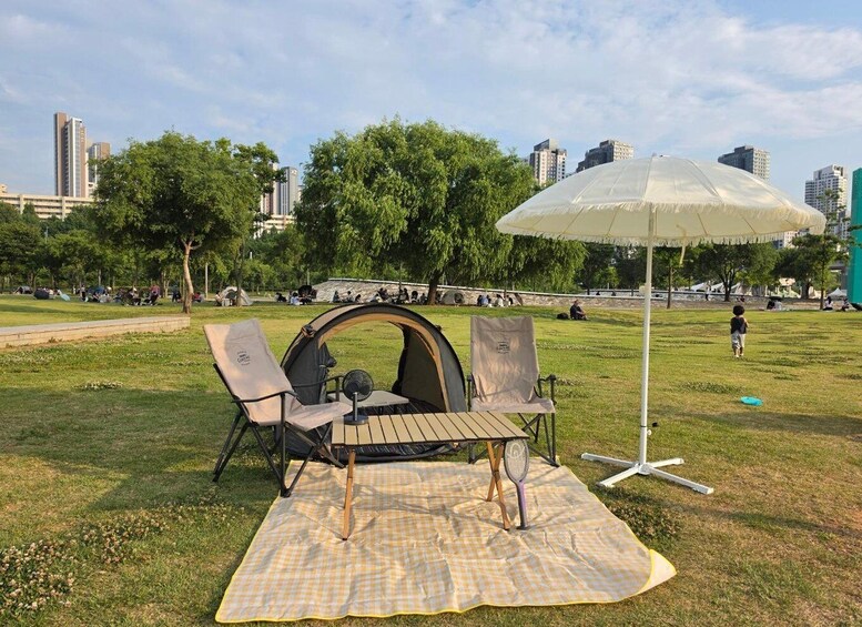 A picnic setup with a tent, chairs, and a table on a grassy field with trees and buildings in the background. 5