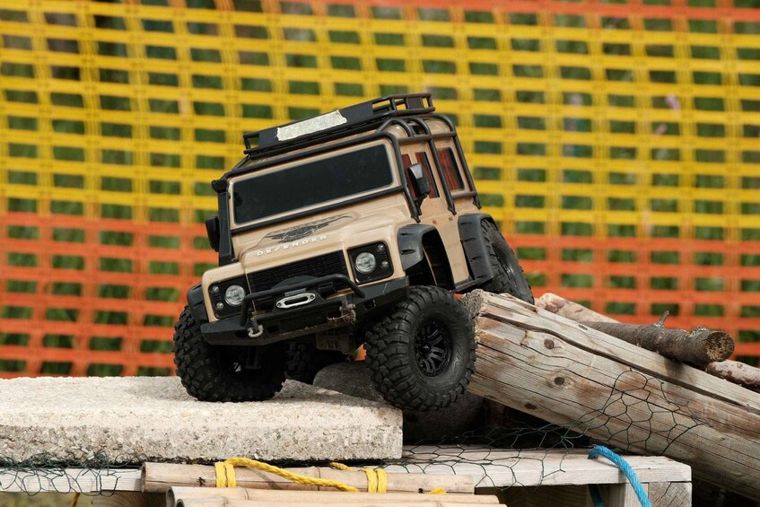 A toy off-road vehicle on a wooden ramp with logs and a wire fence in the background. 5