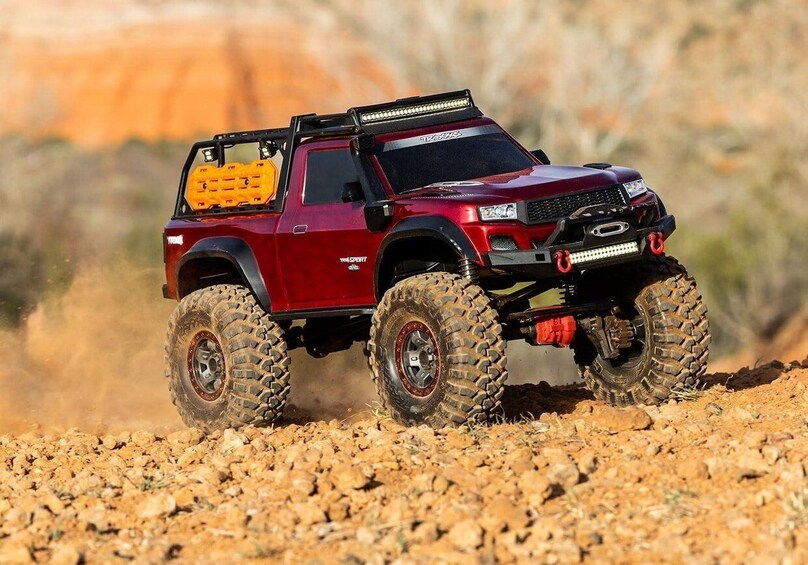 A red off-road vehicle with large tires and a spare tire mounted on the back, driving on a rocky terrain. 2