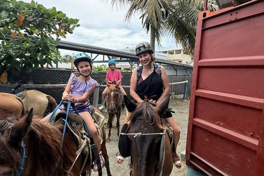 Children riding horses in an outdoor setting with a red trailer and palm trees in the background. 1