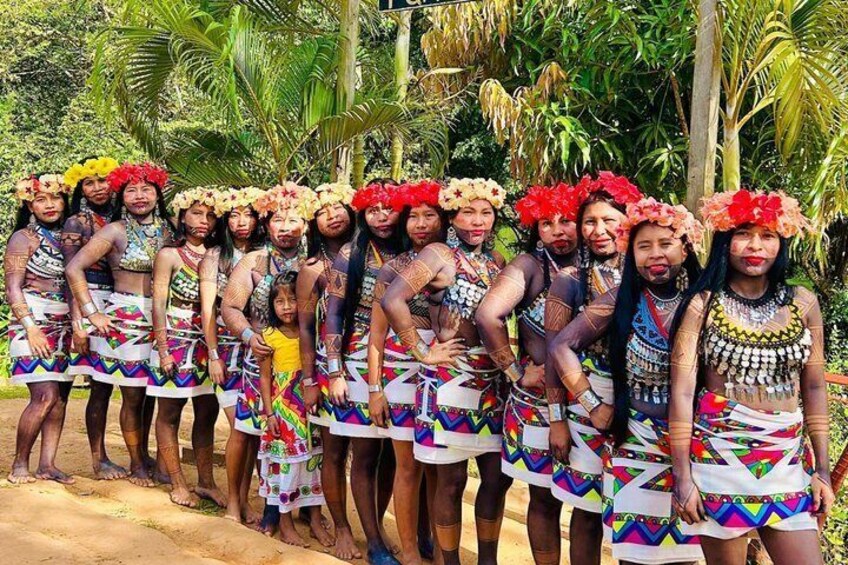 A group of people in traditional attire with floral headdresses and colorful skirts, standing outdoors with greenery in the background. 3