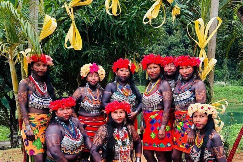 A group of people in traditional attire with elaborate necklaces and headpieces, standing outdoors with greenery in the background. 2
