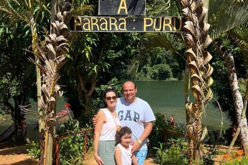 A family posing under a sign with 'A Parara Puru' written on it, surrounded by lush greenery and a body of water. 1