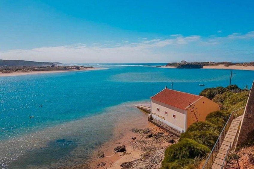 A coastal landscape with a clear blue sea, a sandy beach, and a small white building with a red roof. 2