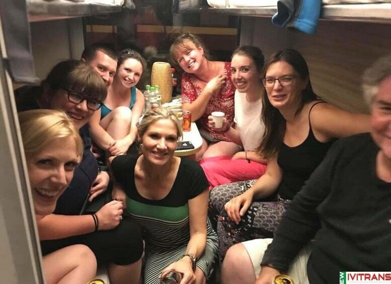 A group of people sitting closely together in a confined space, possibly a train compartment, with a table in the center holding various items. 5