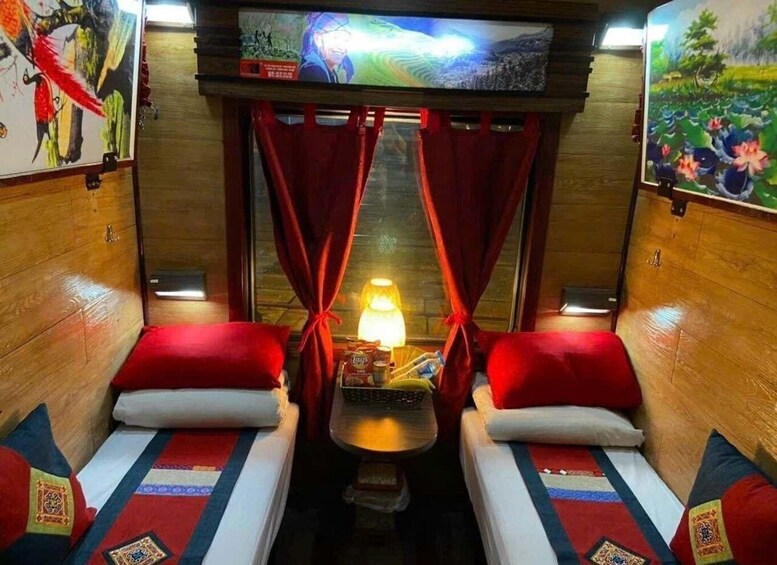 A cozy train compartment with two single beds, red curtains, a table with a lamp, and a painting on the wall. 3
