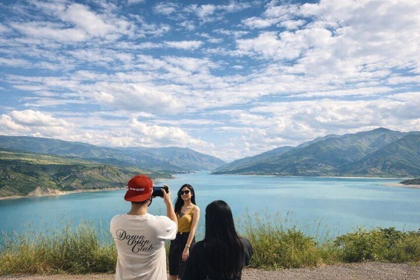 A lake with mountains in the background, a person taking a photo, and two people standing by the lake. 1