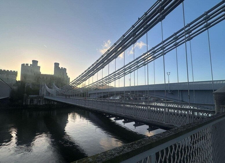 A suspension bridge over a calm river, with a castle in the background. 1