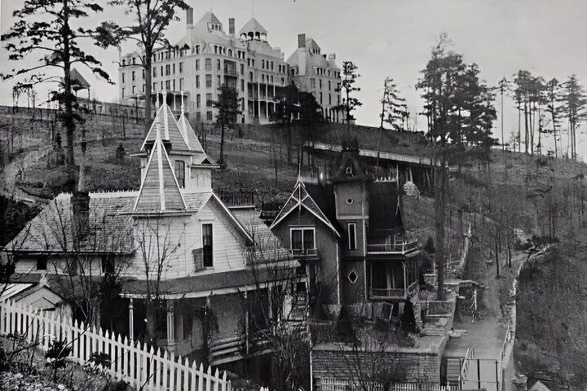 A black and white photograph of a large, multi-story building with a steeply pitched roof, surrounded by trees and a white picket fence. 5