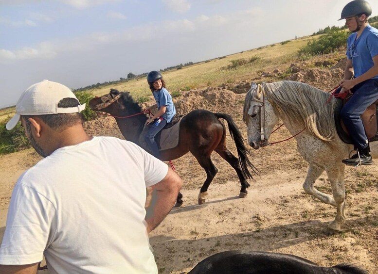 Three people riding horses on a dirt path with sparse vegetation in the background. 1