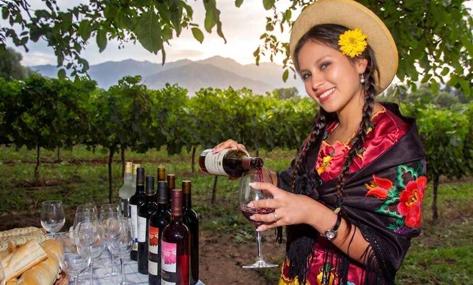 A person pouring wine into a glass, surrounded by wine bottles and glasses, with a vineyard in the background. 1