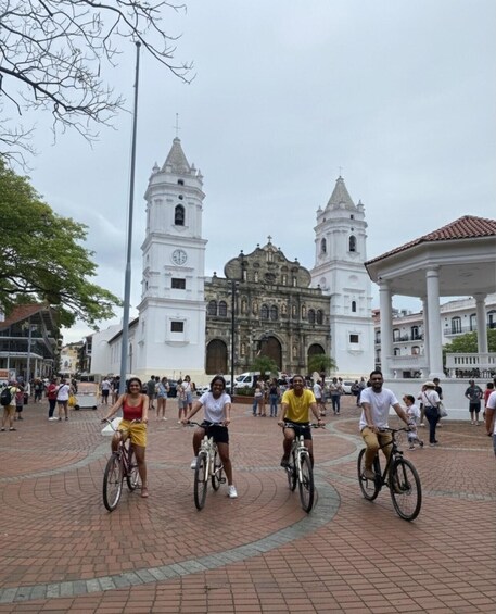 Three people riding bicycles on a paved area with a historic building in the background. 1
