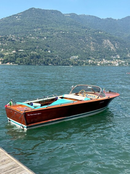 A wooden boat named 'The View Como Lake 2' docked at a pier with a mountainous backdrop. 3