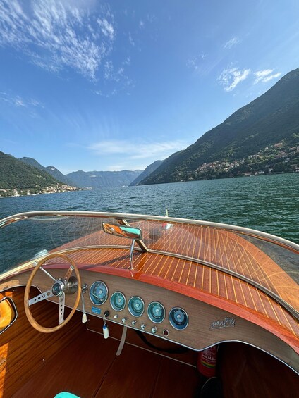A boat's wooden deck with a steering wheel and gauges, surrounded by calm water and a mountainous landscape. 1