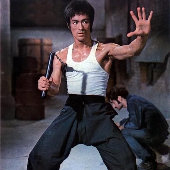 A person in a white tank top and dark pants, holding a gun, with a hand raised in a defensive stance. 1