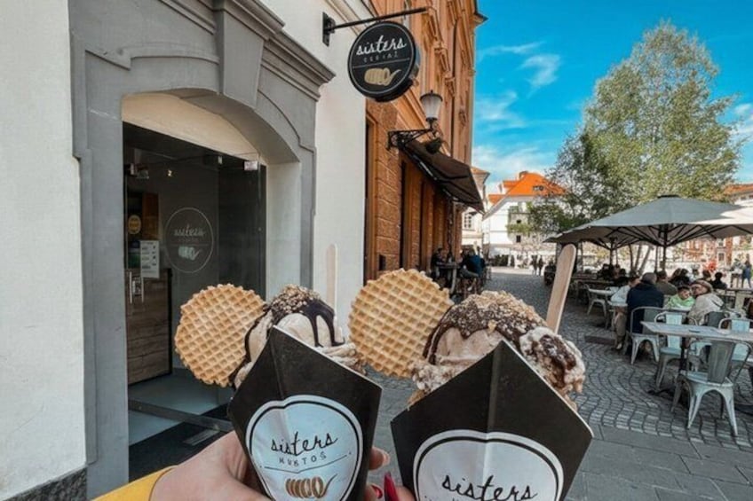 A street with outdoor seating, a building with a sign reading 'sisters,' and waffle cones in the foreground. 2