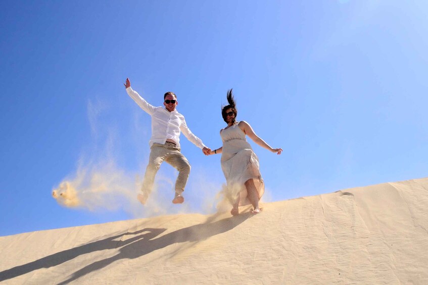 Two people jumping on a sand dune with a clear blue sky. 3