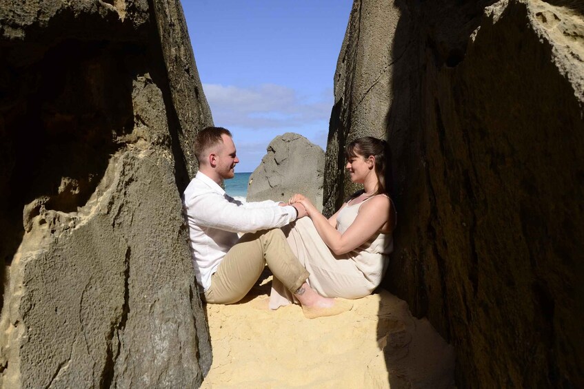 Two people sitting on a sandy beach between rock formations with the ocean in the background. 2