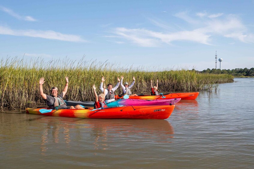 People in colorful kayaks on a calm river, with tall grasses and a clear sky in the background. 1