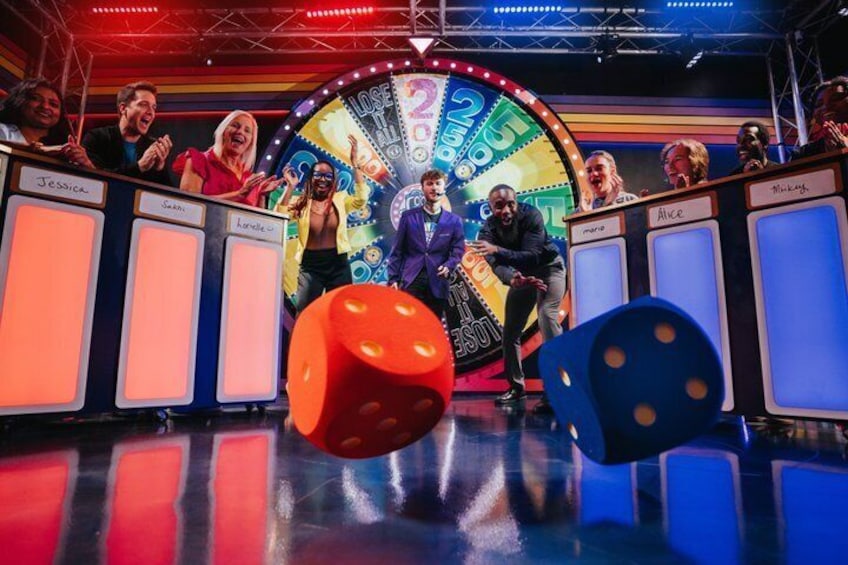 A game show set with a spinning wheel, large dice, and participants seated at a table. 1