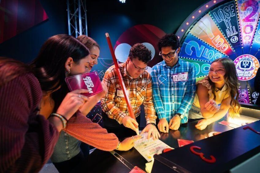 A group of people playing a game with cards and a wheel, with a 'Lose All' sign visible. 2