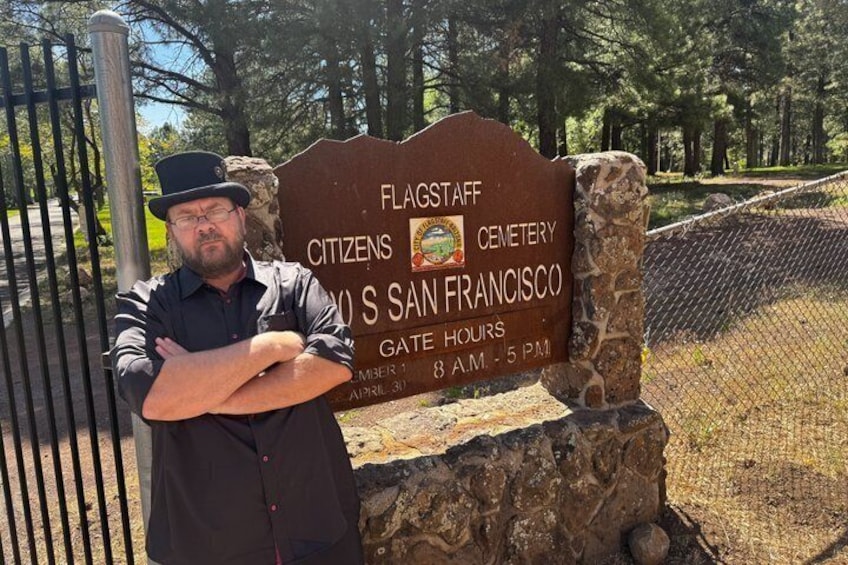 A man standing in front of a sign for Flagstaff Citizens Cemetery in San Francisco with gate hours listed. 1