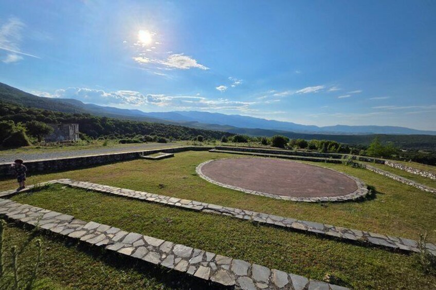 A circular stone structure surrounded by stone walls, set in a grassy area with mountains in the background. 2