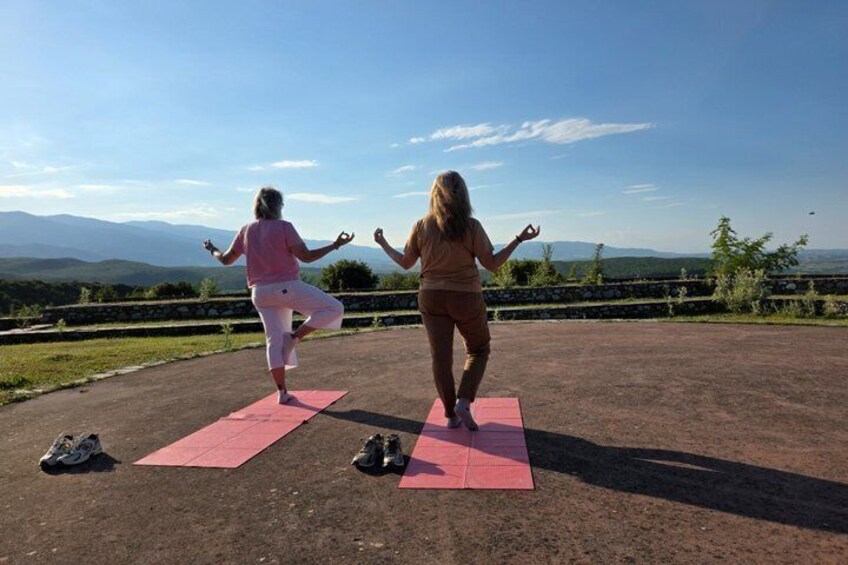 Two people practicing yoga on pink mats outdoors with a scenic view of mountains in the background. 3