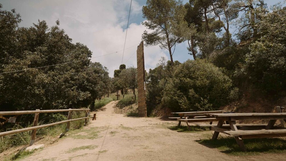 A dirt path flanked by wooden fences and picnic tables, leading to a tall stone monument in a wooded area. 1