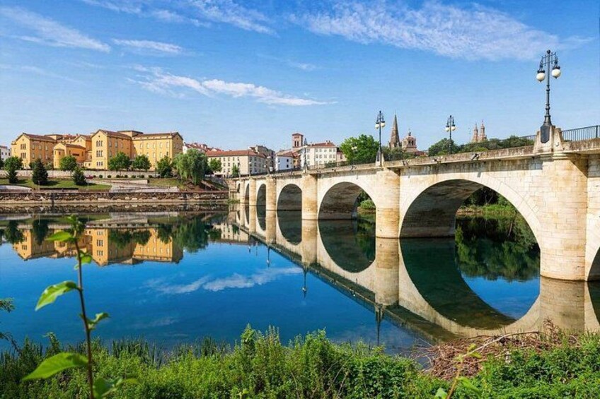 A stone bridge with arches over a calm river, reflecting the surrounding buildings and sky. 4
