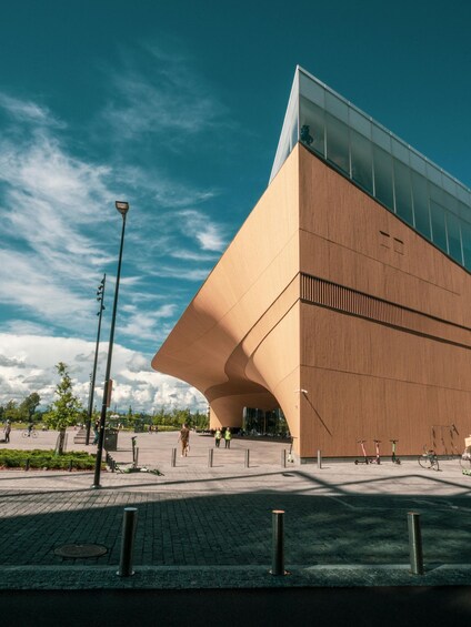 A modern building with a unique curved facade, a clear blue sky, and a paved plaza with bollards and a few people walking. 1