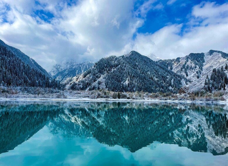 A serene winter landscape with a clear lake reflecting snow-covered mountains and a partly cloudy sky. 5