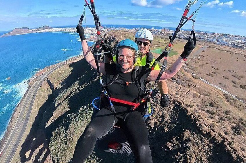 Two people paragliding over a coastal landscape with a road and ocean in the background. 1