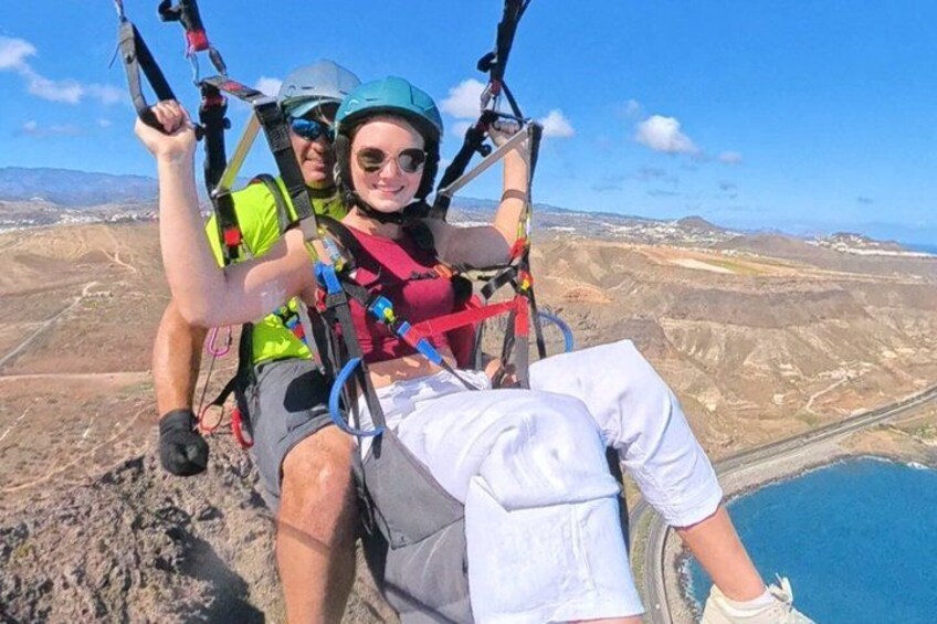 A person in a harness and helmet, seated on a chair, with a mountainous landscape and a body of water in the background. 5