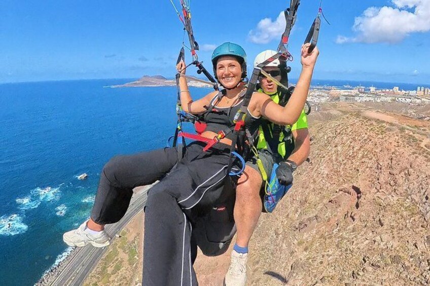 Two people paragliding over a coastal landscape with the ocean and a distant city visible. 1
