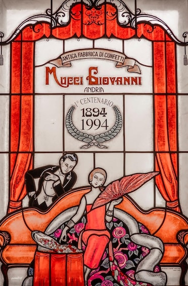 A stained glass window with a couple, a red curtain, and a date marking the 100th anniversary of a confetti factory. 2