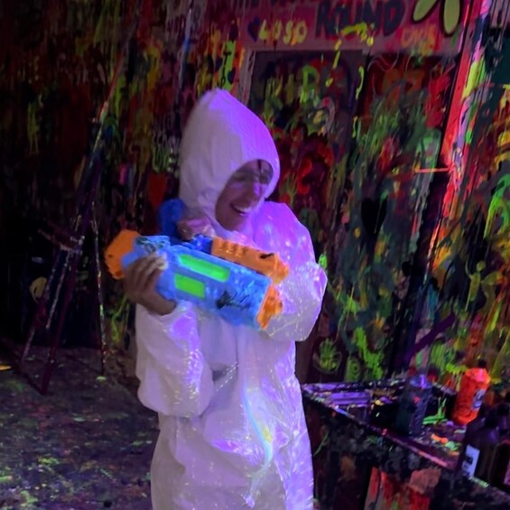 A person in a white hooded garment holding a water gun, with colorful graffiti on the wall behind. 1