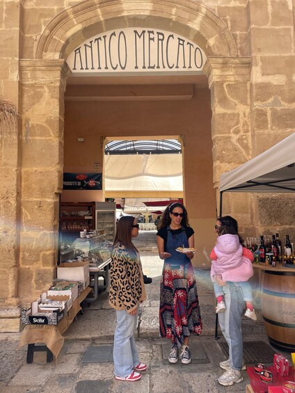 An outdoor market named 'ANTICO MERCATO' with people standing in front, a display of bottled beverages, and a sign for 'Sicilia' coffee. 5