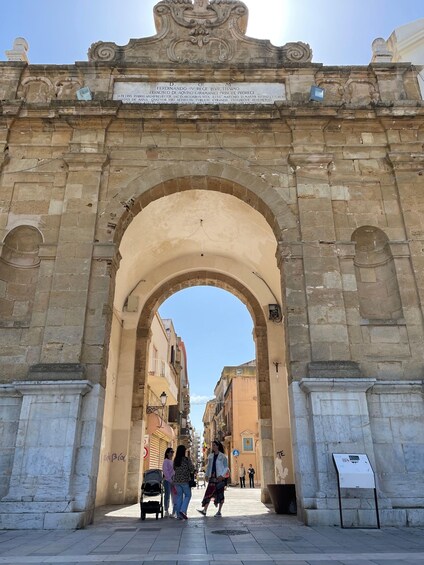 An ornate stone archway with inscriptions, leading into a narrow street lined with buildings. 1