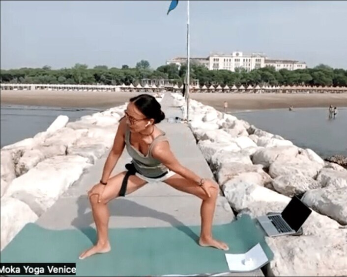 A person performing a yoga pose on a mat by a waterfront with a laptop nearby. 2