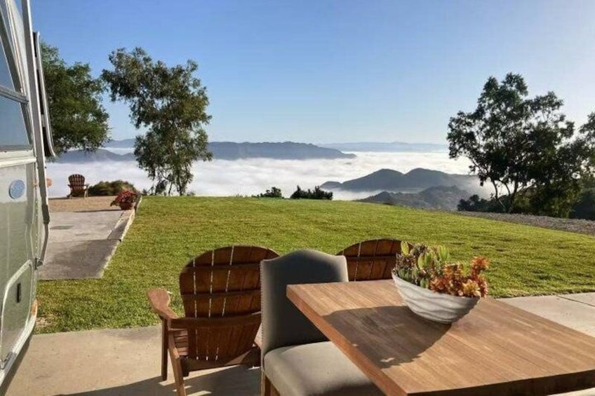 A grassy area with a table and chairs, a wooden bench, and a view of mountains and clouds. 2