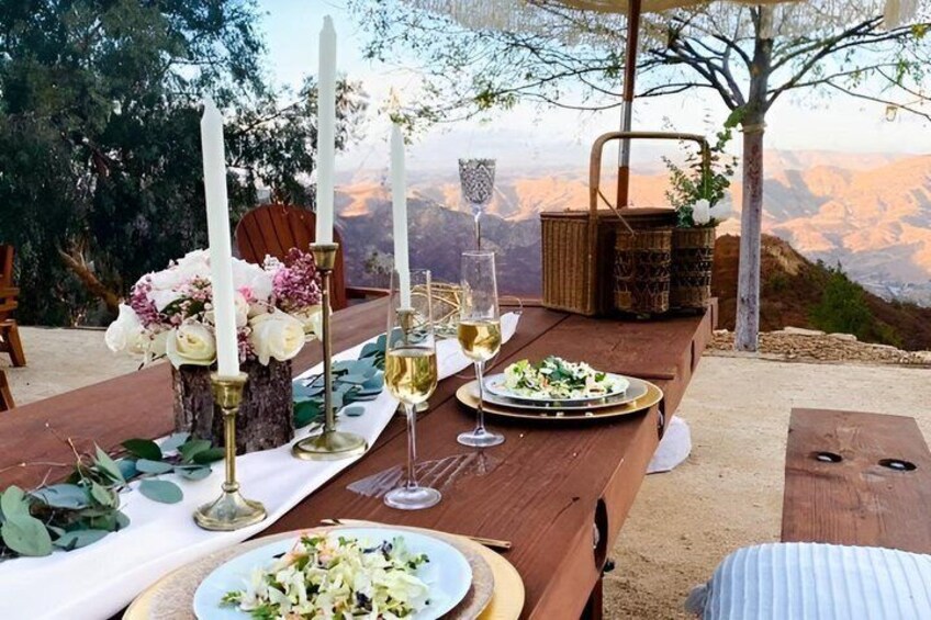 A wooden dining table set outdoors with candles, glasses, and plates of food. 3