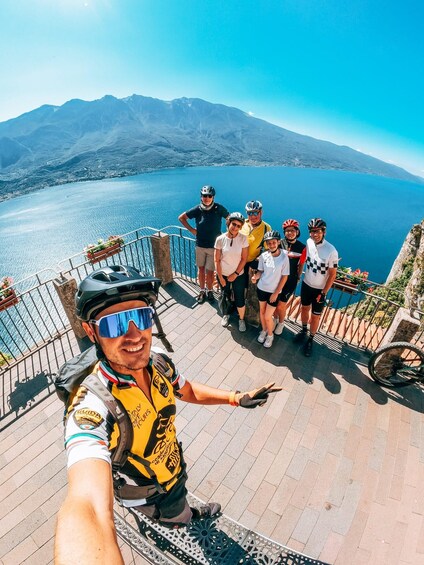 A group of cyclists on a paved path with a scenic view of a lake and mountains in the background. 1