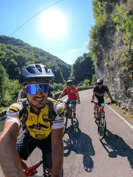 Three cyclists on a paved road with a mountainous backdrop. 2