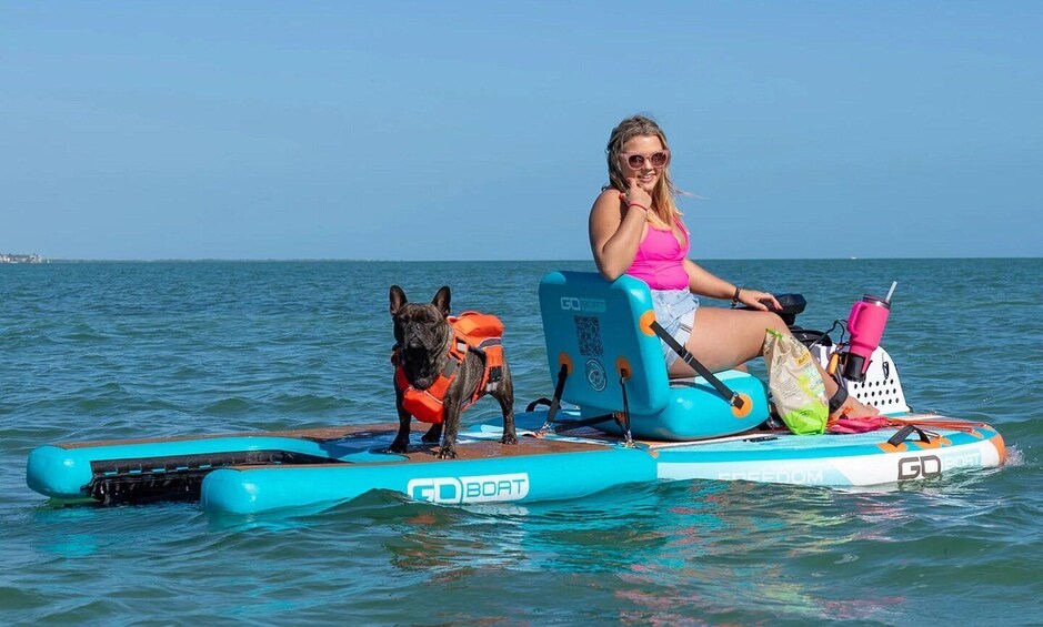 A person on a blue paddleboard with a dog wearing a life jacket, both on calm water. 1