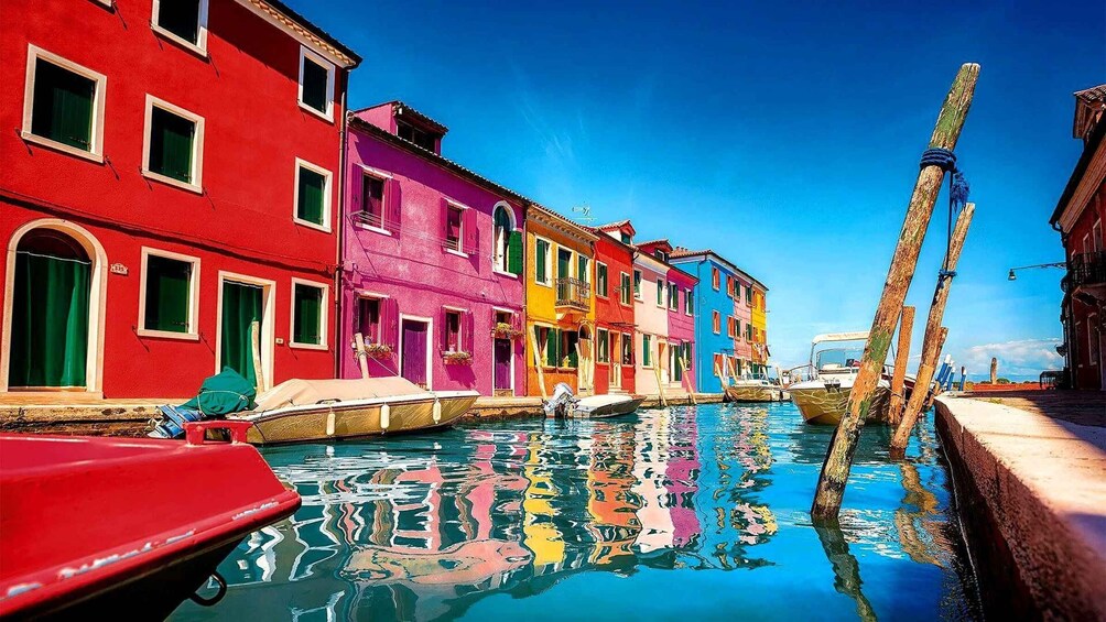 A canal with colorful buildings reflecting in the water, a red boat docked on the left, and wooden poles on the right. 1