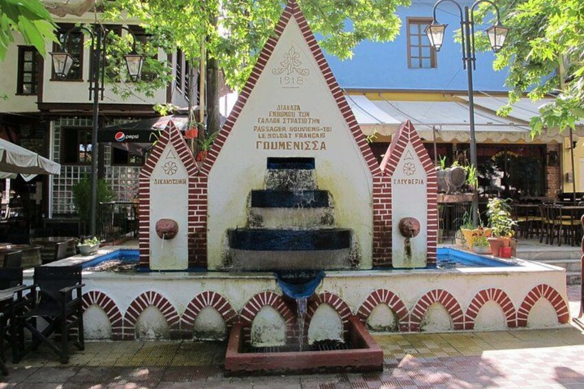 A fountain with inscriptions, surrounded by brick arches and a street cafe in the background. 4