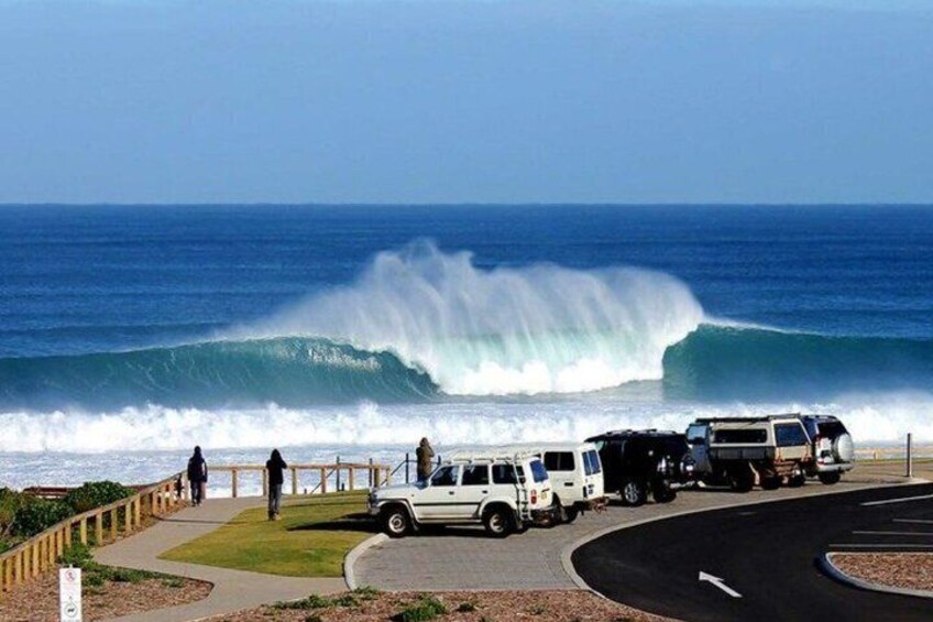 A beach with a large wave, people walking, and parked vehicles. 3